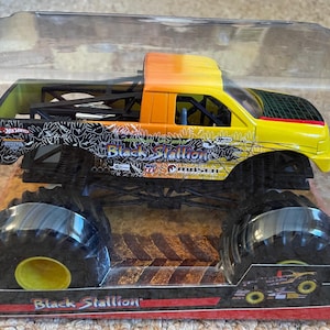 May include: A toy monster truck, the "Black Stallion," is encased in clear plastic. The truck features a yellow and orange cab, black chassis, and oversized black tires with yellow centers. The truck's side displays the name "Black Stallion."