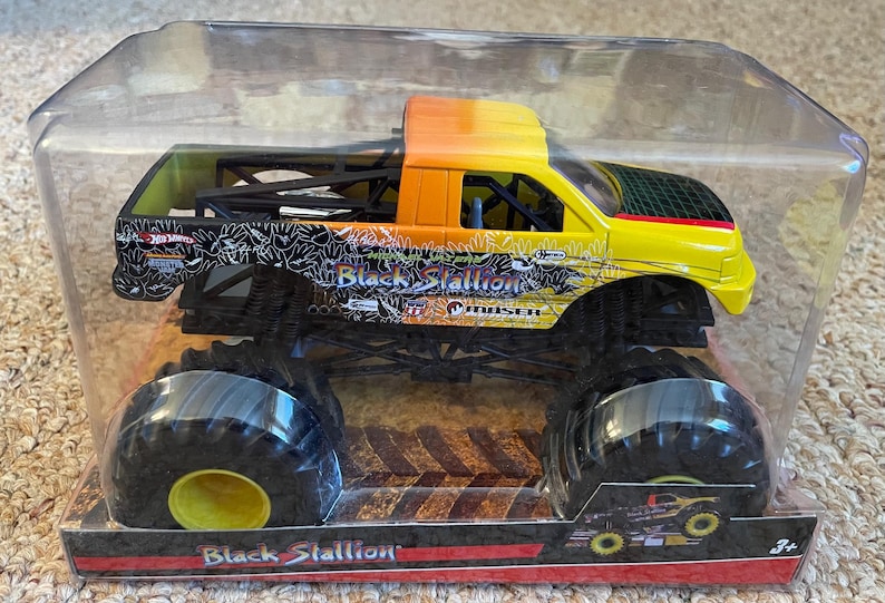May include: A toy monster truck, the "Black Stallion," is encased in clear plastic. The truck features a yellow and orange cab, black chassis, and oversized black tires with yellow centers. The truck's side displays the name "Black Stallion."