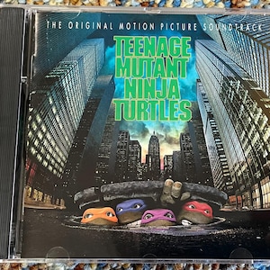 Puede incluir: Caja de CD de la banda sonora de la película Teenage Mutant Ninja Turtles. La portada presenta el título en letras verdes, con las tortugas asomando desde una alcantarilla frente a un paisaje urbano.