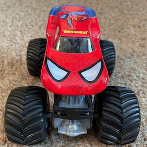 May include: A red and blue Spider-Man monster truck toy with large black tires. The truck features the Spider-Man logo and a web design on the front. The text "SPIDER-MAN" is visible on the side.