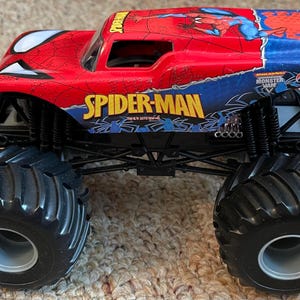 May include: A red, blue, and black Spider-Man monster truck toy. The truck features large black tires, the Spider-Man logo, and a graphic of Spider-Man. The toy is a Hot Wheels Monster Jam vehicle.