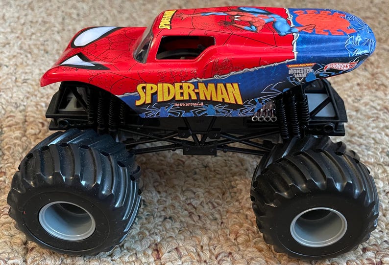 May include: A red, blue, and black Spider-Man monster truck toy. The truck features large black tires, the Spider-Man logo, and a graphic of Spider-Man. The toy is a Hot Wheels Monster Jam vehicle.