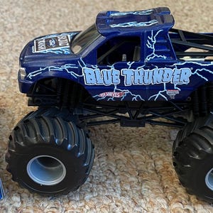 May include: A blue toy monster truck with the words "BLUE THUNDER" and lightning bolt graphics. The truck has large black tyres and a "BUILT TOUGH" sticker. A partially visible box with "MONSTER JAM" branding is on the left.