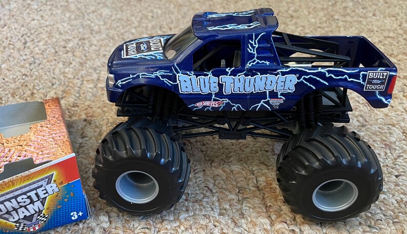 May include: A blue toy monster truck with the words "BLUE THUNDER" and lightning bolt graphics. The truck has large black tyres and a "BUILT TOUGH" sticker. A partially visible box with "MONSTER JAM" branding is on the left.