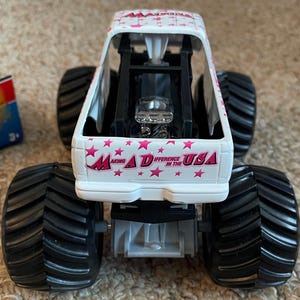 K&ouml;nnte beinhalten: Ein wei&szlig;-rosa Spielzeug-Monstertruck mit schwarzen Reifen. Der Truck tr&auml;gt den Text "Making a Difference in the USA" und rosa Sterne. Der Truck hat einen detaillierten Motor und einen &Uuml;berrollk&auml;fig.