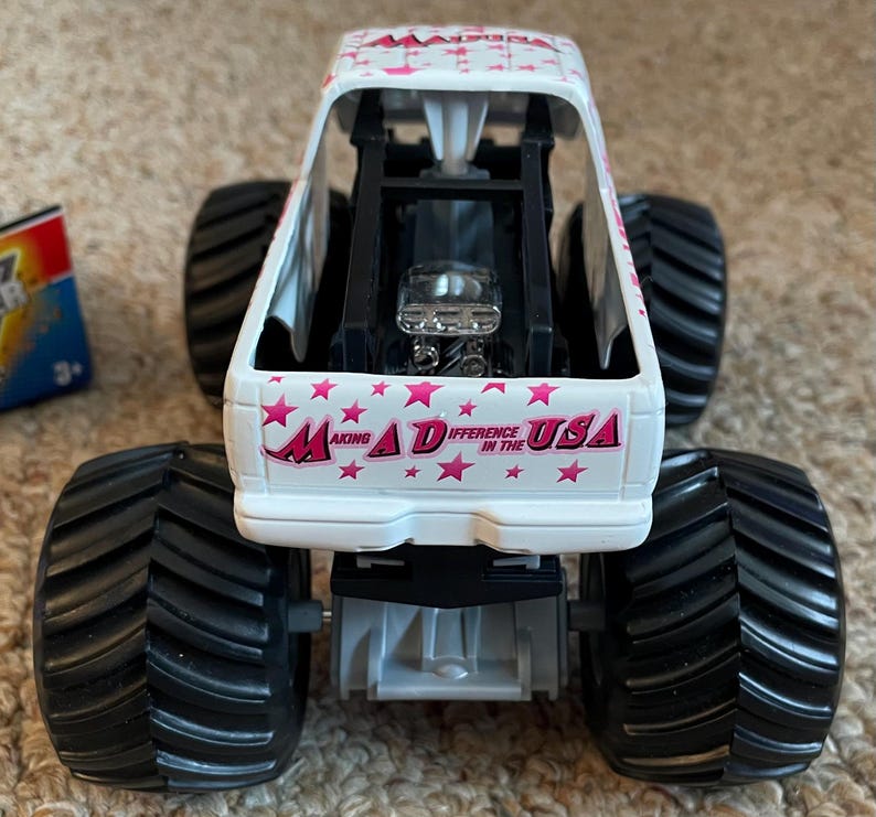 K&ouml;nnte beinhalten: Ein wei&szlig;-rosa Spielzeug-Monstertruck mit schwarzen Reifen. Der Truck tr&auml;gt den Text "Making a Difference in the USA" und rosa Sterne. Der Truck hat einen detaillierten Motor und einen &Uuml;berrollk&auml;fig.