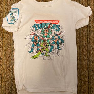 Puede incluir: Camiseta blanca con un diseño de las Tortugas Ninja. El diseño muestra a las cuatro tortugas en acción, con las palabras "Teenage Mutant Ninja Turtles" encima. Un parche de "Mutant Ninja Turtles" está en la manga izquierda.