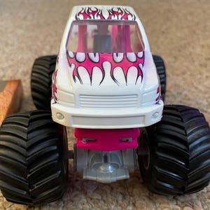 May include: A white toy monster truck with pink flame graphics on the hood and roof. The truck has large black tires and pink accents. The front of the truck is visible, showcasing the grill and headlights.