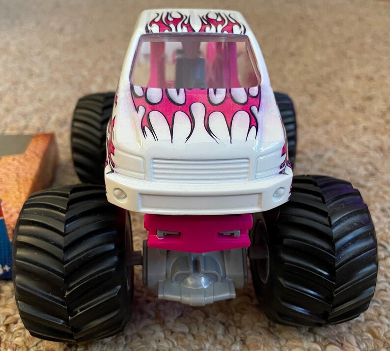 May include: A white toy monster truck with pink flame graphics on the hood and roof. The truck has large black tires and pink accents. The front of the truck is visible, showcasing the grill and headlights.