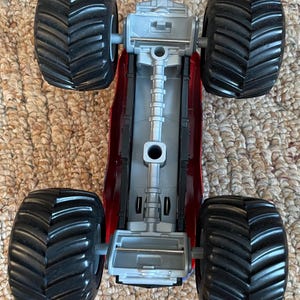 May include: A red and gray toy monster truck with four large black tires. The truck's chassis is red, and the engine components are gray. The tires have a deep tread pattern.