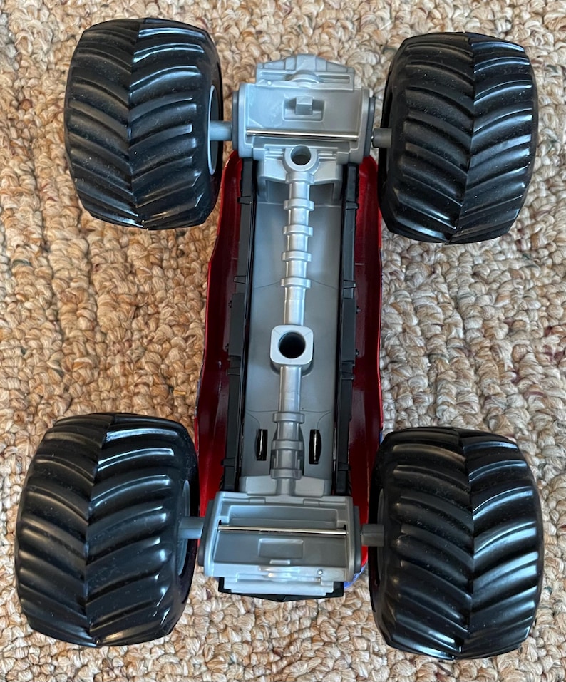 May include: A red and gray toy monster truck with four large black tires. The truck's chassis is red, and the engine components are gray. The tires have a deep tread pattern.