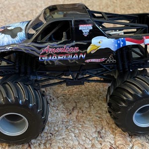 May include: Black toy monster truck with large black tires. The truck features patriotic eagle graphics and the words "American Guardian." The truck has a black roll cage and the Hot Wheels logo.