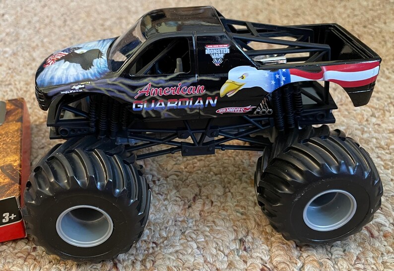 May include: Black toy monster truck with large black tires. The truck features patriotic eagle graphics and the words "American Guardian." The truck has a black roll cage and the Hot Wheels logo.