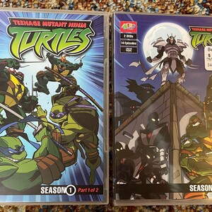 Op de afbeelding: Twee dvd-hoesjes van Teenage Mutant Ninja Turtles Seizoen 1, Deel 1 en Deel 2. De covers tonen actiescènes van de Turtles in gevecht, met de serietitel in groen en geel. De Deel 2 hoes heeft een Best Buy sticker.