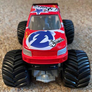 May include: A red toy monster truck with large black tyres. The truck features a white and blue logo on the front and the words "Nitro Circus". The truck has a silver undercarriage and a red cab.