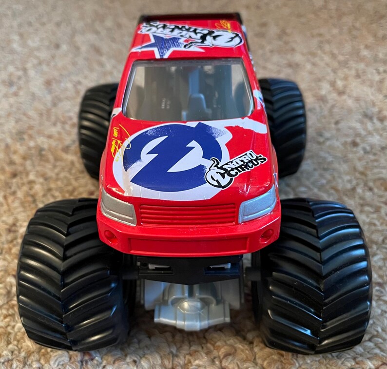 May include: A red toy monster truck with large black tyres. The truck features a white and blue logo on the front and the words "Nitro Circus". The truck has a silver undercarriage and a red cab.