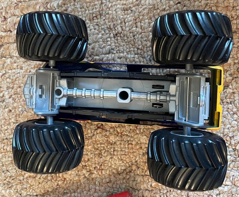 Może przedstawiać: Zabawkowy monster truck z czterema dużymi czarnymi oponami i ż&oacute;łto-niebieskim podwoziem. Widoczna jest dolna strona ciężar&oacute;wki, pokazująca osie i szczeg&oacute;ły zawieszenia. Opony mają głębokie bieżniki zapewniające przyczepność w terenie.
