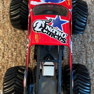 May include: A red toy monster truck with black tyres. The truck features the "Nitro Circus" logo and a blue lightning bolt design. The tyres are large and have a deep tread pattern.