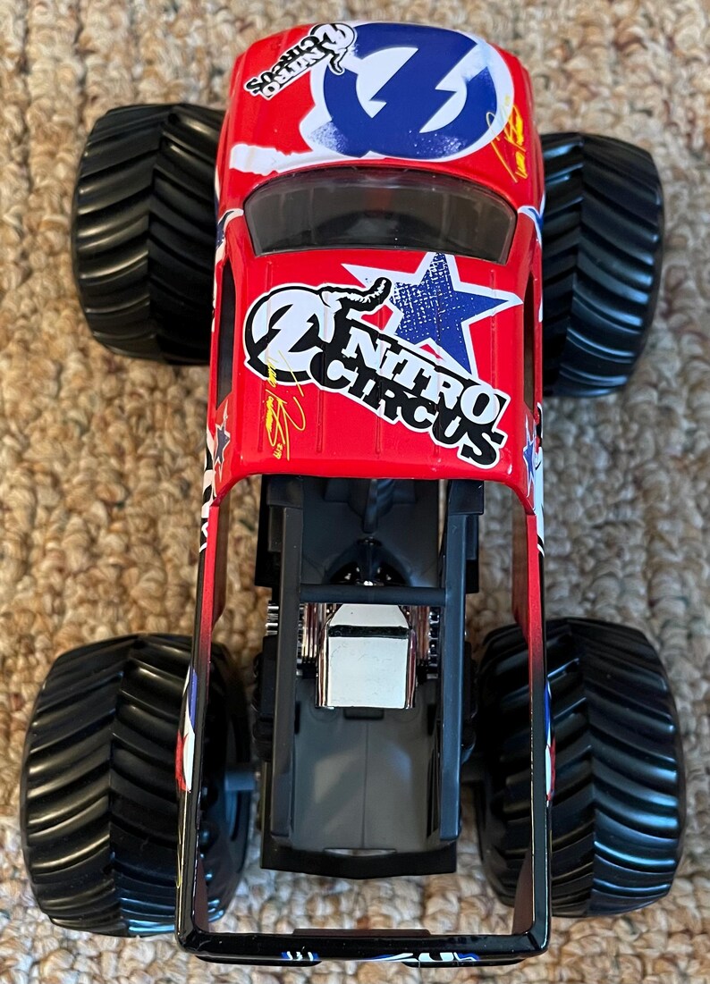May include: A red toy monster truck with black tyres. The truck features the "Nitro Circus" logo and a blue lightning bolt design. The tyres are large and have a deep tread pattern.