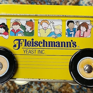 May include: A yellow rectangular metal tin shaped like a bus with black wheels. The side features illustrations of children in windows and the text Fleischmann's YEAST INC. in blue lettering. The design includes a driver and various passengers.
