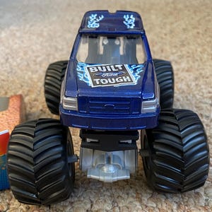 May include: A blue toy monster truck with large black tyres. The truck has the words "BUILT Ford TOUGH" on the hood. The truck is a toy vehicle.