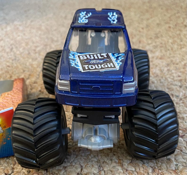 May include: A blue toy monster truck with large black tyres. The truck has the words "BUILT Ford TOUGH" on the hood. The truck is a toy vehicle.