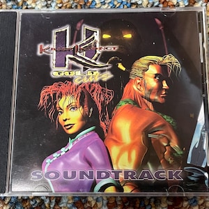 Puede incluir: Caja de CD de la banda sonora de Killer Instinct Gold Cuts. La portada presenta dos personajes ilustrados, una mujer de morado y un hombre de naranja, con el logo del juego en morado y dorado. La palabra "SOUNDTRACK" está impresa en blanco.