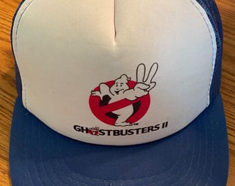 Op de afbeelding: Een blauw-witte Ghostbusters II truckerpet. De pet heeft een wit voorpaneel met het Ghostbusters-logo in rood en wit. De klep en de mesh-achterkant zijn blauw.