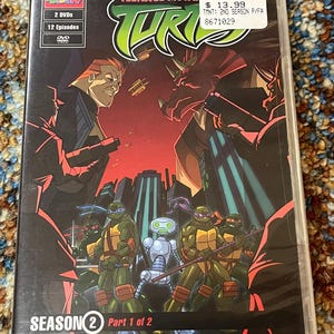 Op de afbeelding: Teenage Mutant Ninja Turtles Seizoen 2 Deel 1 van 2 dvd-set. De cover toont de Turtles, een robot en schurken in een stadsomgeving. De dvd-hoes heeft een zwarte achtergrond met groene, rode en gele accenten.