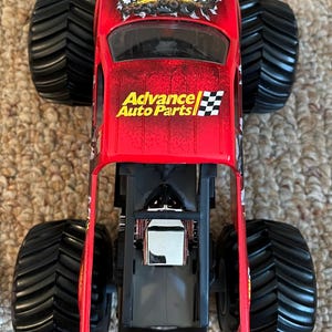 May include: Red toy monster truck with black tires. The truck has the word "Grinder" in a distressed font on the roof and "Advance Auto Parts" on the side. The truck has a detailed engine compartment.