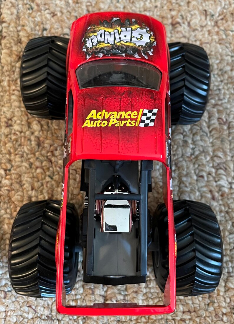 May include: Red toy monster truck with black tires. The truck has the word "Grinder" in a distressed font on the roof and "Advance Auto Parts" on the side. The truck has a detailed engine compartment.