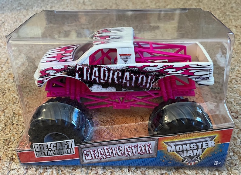 May include: Die-cast metal body toy monster truck, the "Eradicator", in a clear plastic package. The truck is white with pink and black flame graphics, pink chassis, and large black tires. The text "Eradicator" is visible on the side.