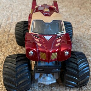 May include: A red and gold toy monster truck with large black tires. The truck has a gold front section and a red body with gold accents. The tires are thick and have a deep tread pattern. The truck is designed to look like a superhero.