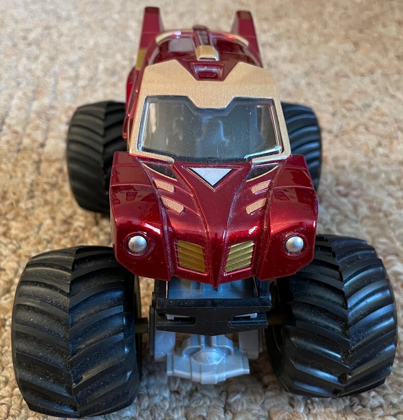 May include: A red and gold toy monster truck with large black tires. The truck has a gold front section and a red body with gold accents. The tires are thick and have a deep tread pattern. The truck is designed to look like a superhero.