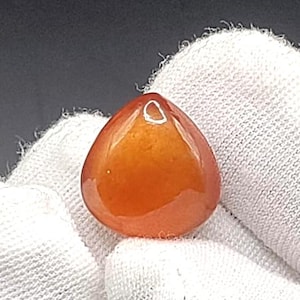 May include: A polished, teardrop-shaped gemstone in shades of orange and amber. The stone has a smooth, glossy surface and a small, white-rimmed hole at the top. The gem is held against a white, textured fabric.