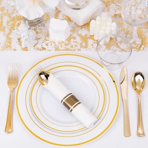 Puede incluir: Un juego de platos de plástico blanco con borde dorado. Los platos están apilados uno encima del otro. También hay cubiertos de plástico dorado sobre una superficie blanca.