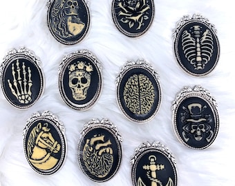 May include: Ten black and white enamel pins with silver frames. Each pin features a different image, including a skull, a rose, a brain, a heart, an anchor, a skeleton, a horse, and a hand.