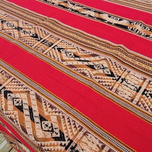 May include: A red woven textile with geometric patterns in tan, black, and brown. The textile features wide red stripes and detailed borders with repeating designs. The overall design is symmetrical and the textile appears to be made of a thick, durable material.