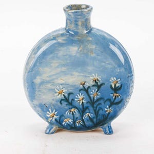 May include: A round, blue ceramic vase with a short neck and three small feet. The vase is decorated with a floral design of white daisies with yellow centers and green stems. The background has a cloudy effect.