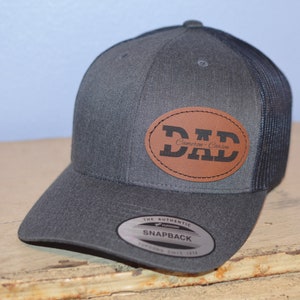 May include: A gray and black trucker hat with a brown leather patch that says "DAD" with the text "Cameron - Carson" underneath. The hat has a black mesh back and a gray front.