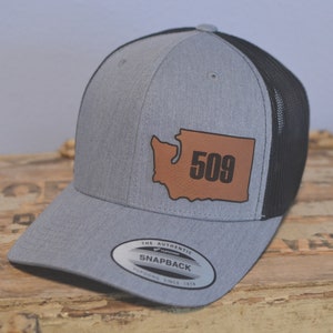 May include: Gray and black Snapback hat with a brown leather patch featuring the outline of Washington state and the number 509. The hat has a mesh back and a curved brim. The text "THE AUTHENTIC SNAPBACK YUPOONG SINCE 1974" is on the front.