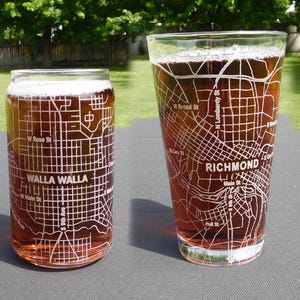 May include: Two clear glass drinking vessels filled with amber liquid. The left is a can-shaped glass etched with a Walla Walla street map. The right is a tapered pint glass etched with a Richmond street map, including labels for W Broad St and Main St.