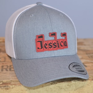 May include: A gray and white trucker hat with a red and black castle-shaped patch that says "Jessica" on it. The hat has a white mesh back and a gray front.