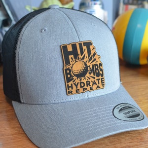 May include: A gray and black trucker hat with a brown leather patch that says "Hit Bombs Hydrate Repeat" with a golf ball graphic. The hat has a black mesh back and a gray front.