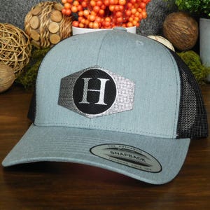 May include: A light blue trucker hat with a black mesh back. The front features a silver and black emblem with the letter "H". The hat has a curved brim and a snapback closure.