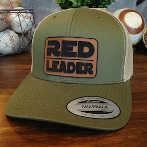 May include: A green and beige baseball cap with a brown leather patch that says "RED LEADER" in black letters. The cap has a mesh back and a snap closure.