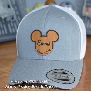 May include: A gray and white Snapback hat with a wood grain Mickey Mouse patch. The patch features the name "Emma" and the text "Disney 2024". The hat has a curved brim and a snap closure.