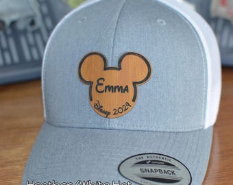 May include: A gray and white Snapback hat with a wood grain Mickey Mouse patch. The patch features the name "Emma" and the text "Disney 2024". The hat has a curved brim and a snap closure.