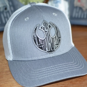 May include: A gray and white baseball cap with a black and silver Rebel Alliance symbol on the front. The symbol is made up of a circle with a stylized design inside.