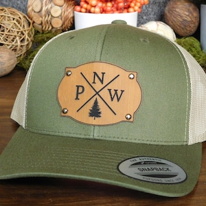 May include: Olive green and tan trucker hat with a leather patch. The patch features the letters "PNW" with a tree graphic. The hat has a curved brim and a snapback closure. The hat is made of a durable material.